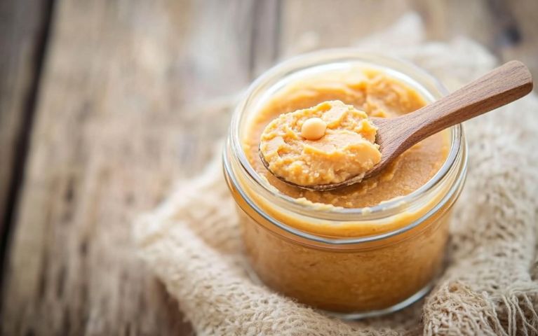 Miso di ceci cremoso in un barattolo di vetro, presentato su un tavolo di legno chiaro con un cucchiaio di legno che ne mostra la consistenza